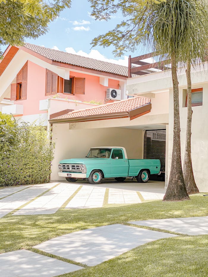 A teal vintage pickup truck parked in a suburban garage under a sunny sky, surrounded by trees and a neat lawn.